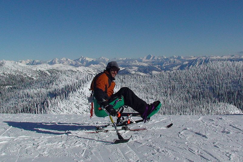 person in a monoski on a mountain summit with mountain range and wide sky in the background